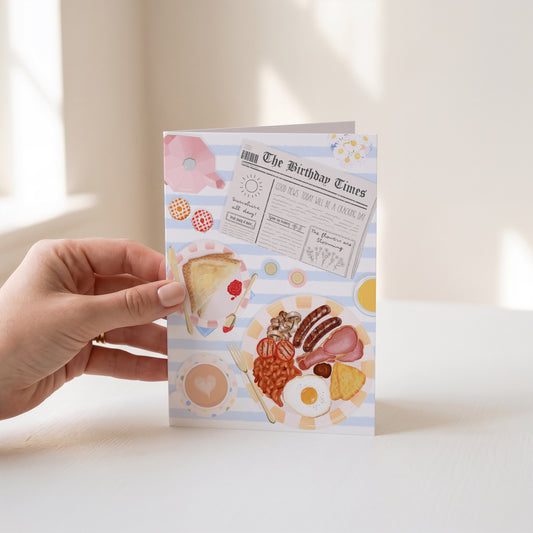 A hand holds a greeting card featuring a vintage breakfast illustration with a newspaper, toast, eggs, beans, bacon, and a cup of tea against a light blue and white striped background.