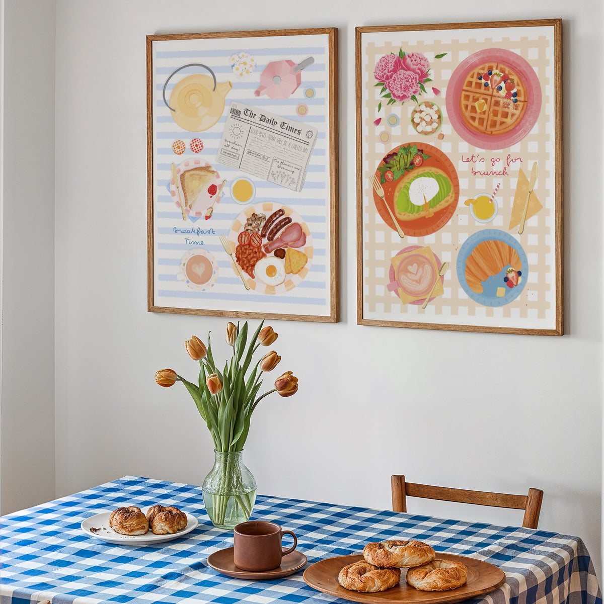 Two framed breakfast-themed art prints hanging on a white wall above a blue-and-white checkered table with pastries, a mug, and tulips.
