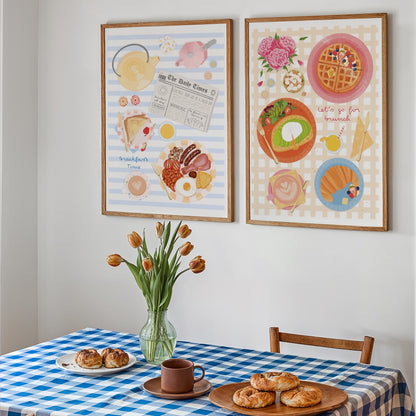 Two framed breakfast-themed art prints hanging on a white wall above a blue-and-white checkered table with pastries, a mug, and tulips.
