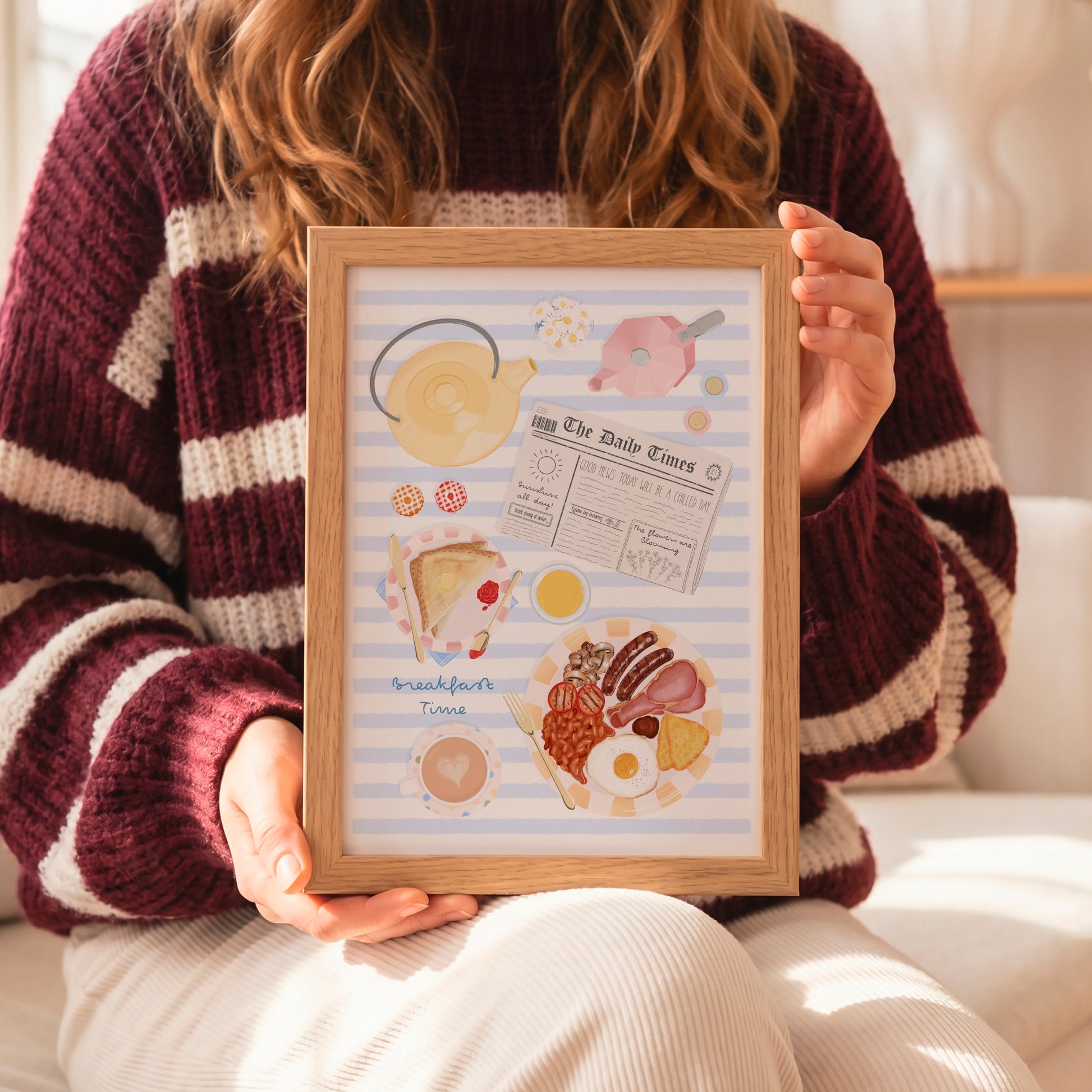 A person in a burgundy and cream striped knit sweater holds a framed hand-drawn art print featuring breakfast illustrations like a teapot, newspaper, toast, and eggs on a light blue striped background.