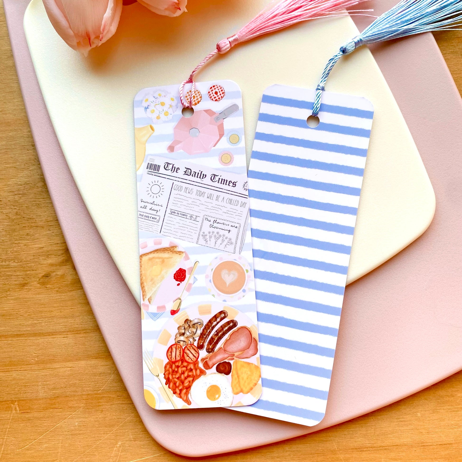Two colourful blue striped bookmarks with breakfast food illustrations on a pink surface with a wooden background