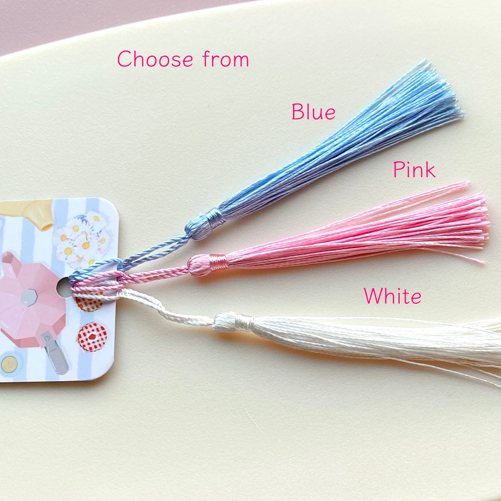 Three bookmark tassels in blue, pink, and white with labels indicating colour options on a light background.