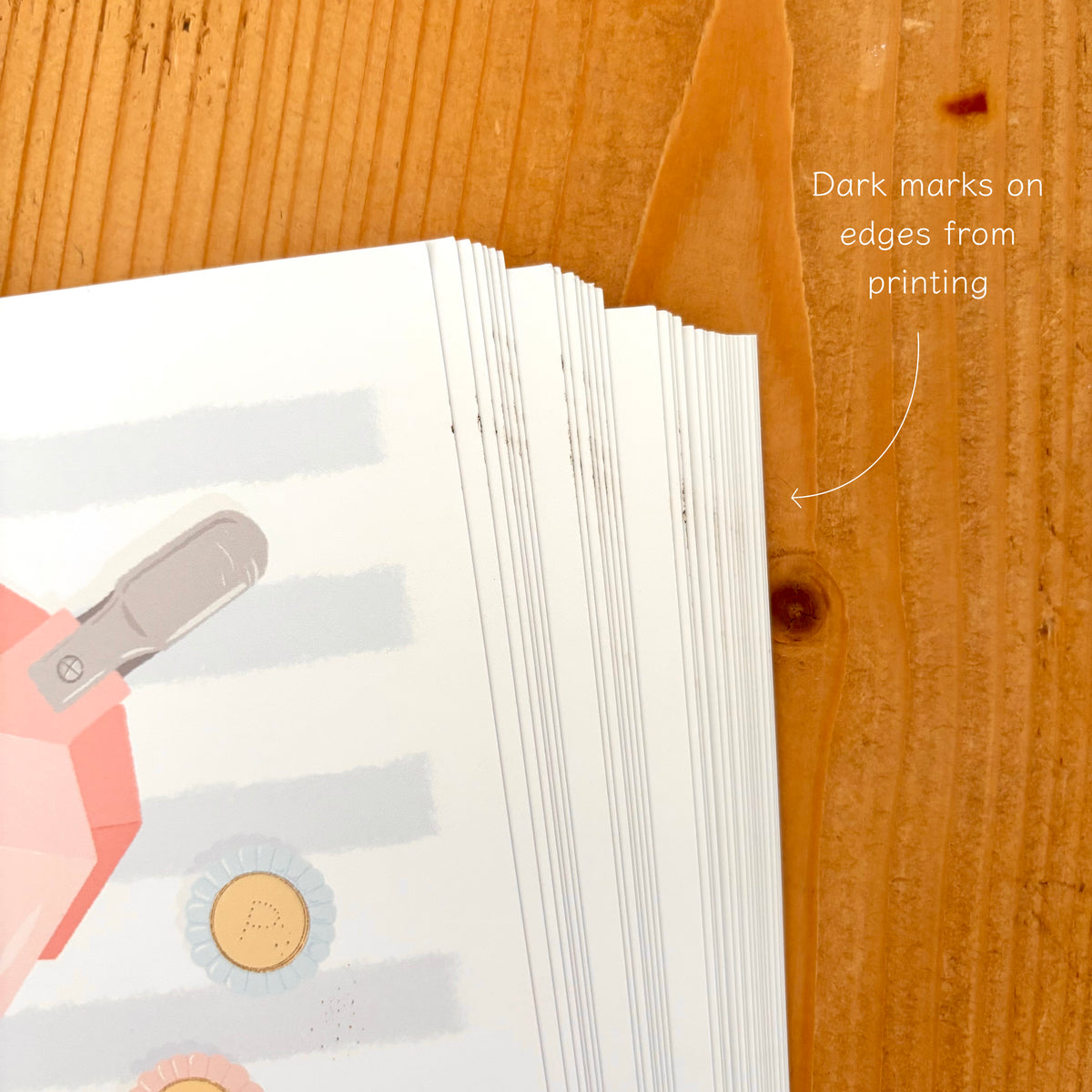 A close-up of a stack of printed paper sheets on a wooden surface, with a handwritten note pointing to dark marks on the edges from printing.