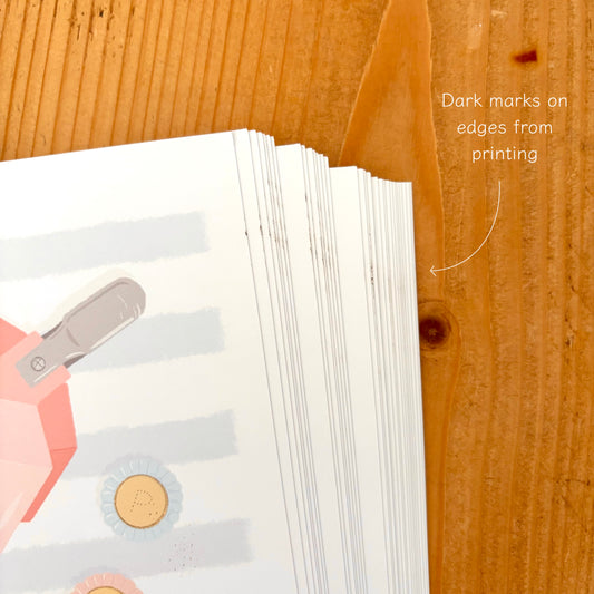 A close-up of a stack of printed paper sheets on a wooden surface, with a handwritten note pointing to dark marks on the edges from printing.