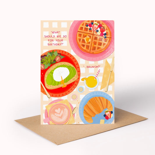 A birthday greeting card with a whimsical breakfast food illustration, shown standing upright on a brown kraft envelope against a clean white background.