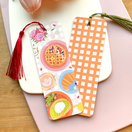 Two decorative bookmarks illustrated with brunch food with tassels on a wooden surface
