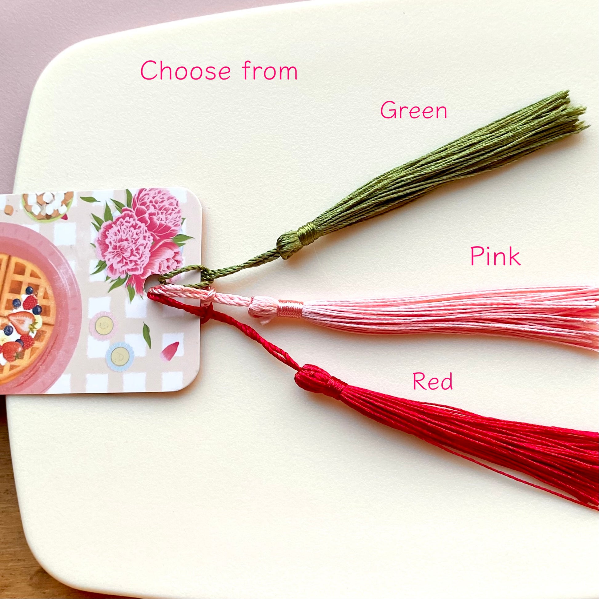 Three bookmark tassels in green, pink, and red on a white surface.