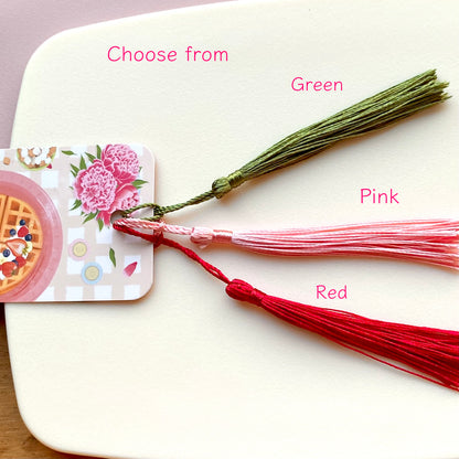 Three bookmark tassels in green, pink, and red on a white surface.