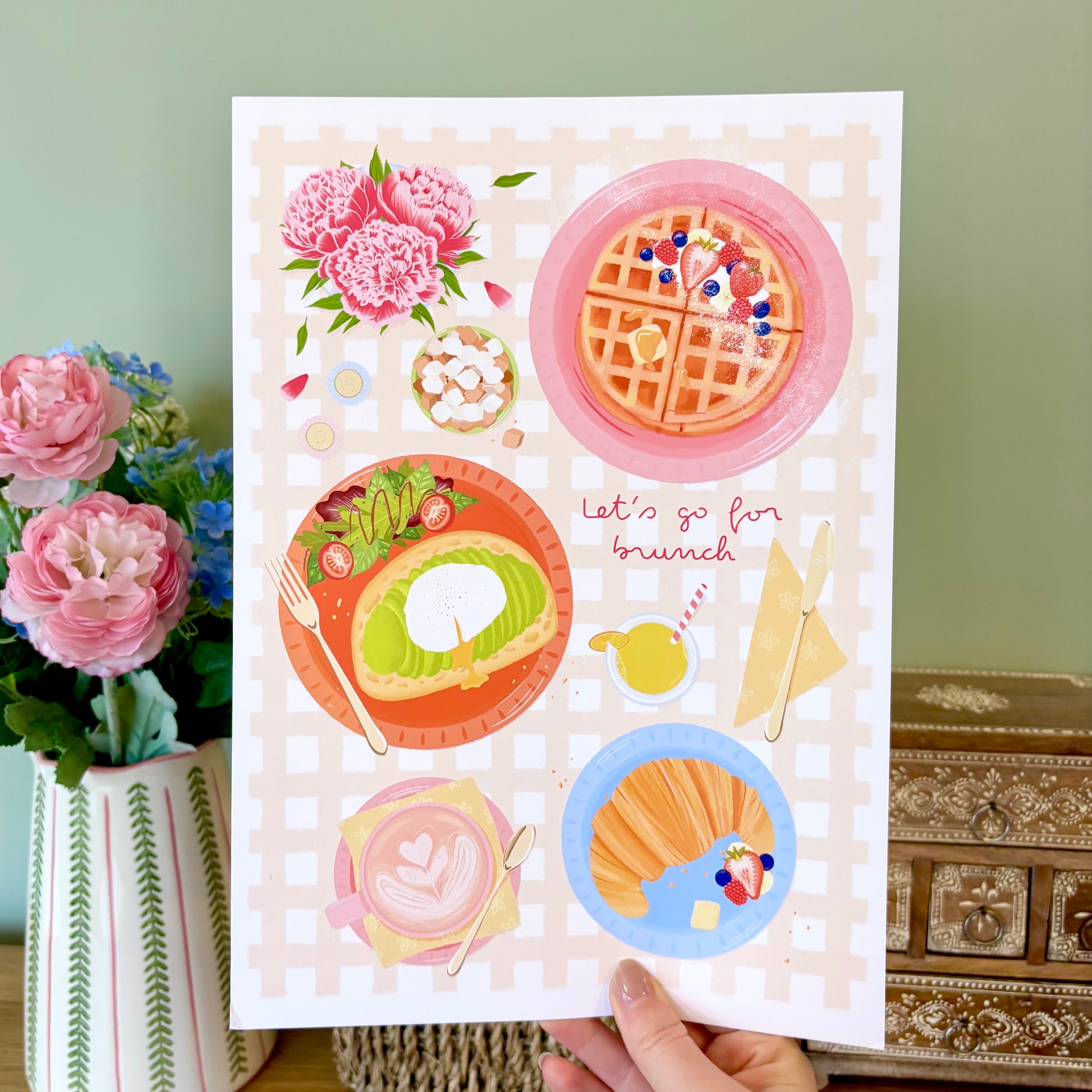Art print with illustrations of brunch food and flowers held by a hand, with a decorative background.