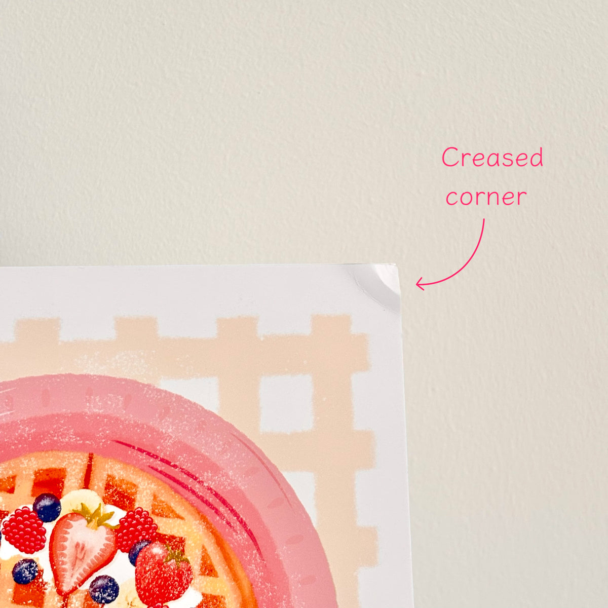 A close-up of a printed sheet showing a pink waffle with berries and a cream grid pattern, with a hand peeled back the top-right corner to demonstrate a creased corner. Pink text reads 'Creased corner' on a light beige wall.