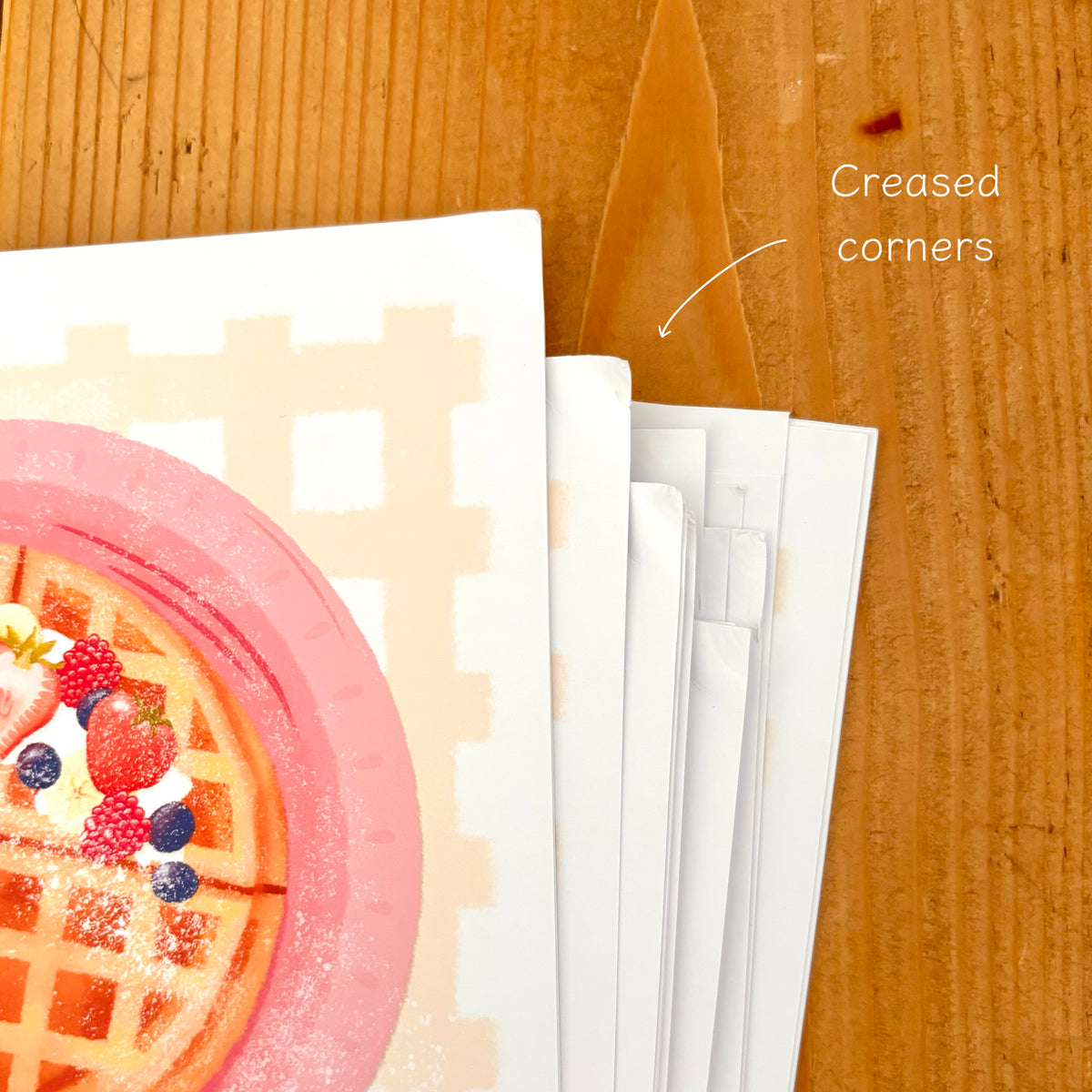 A stack of white paper prints with a printed waffle illustration on one sheet, shown on a wooden surface. White text reads 'Creased corners' with an arrow pointing to the paper.
