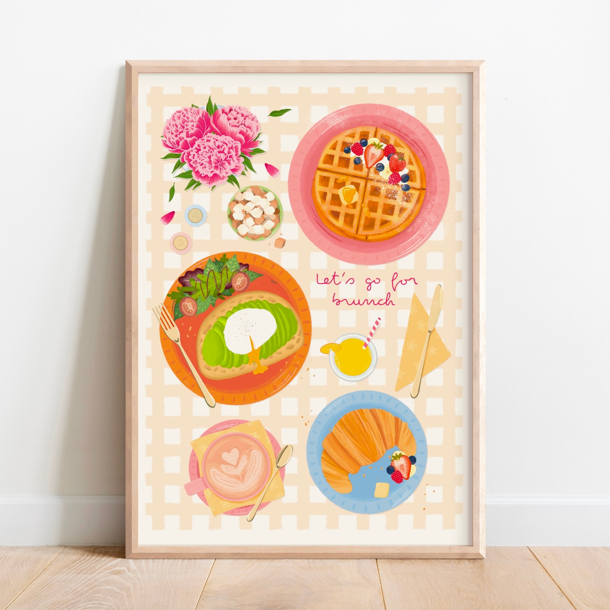 A framed art print featuring a whimsical breakfast illustration with waffles, avocado toast, a croissant with berries, and pink flowers, leaning against a white wall on a light wood floor.