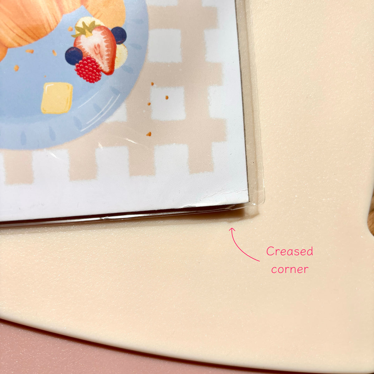 Close-up of a cream-coloured acrylic or plastic tray corner with a pink arrow and text reading 'Creased corner,' showing a printed card partially tucked into the edge.