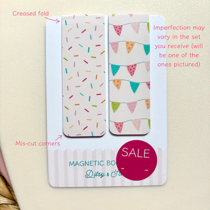 A set of two decorative magnetic bookmarks on a white backing card, photographed against a light cream background. One bookmark has multicoloured confetti on a white base, and the other has colourful bunting flags.