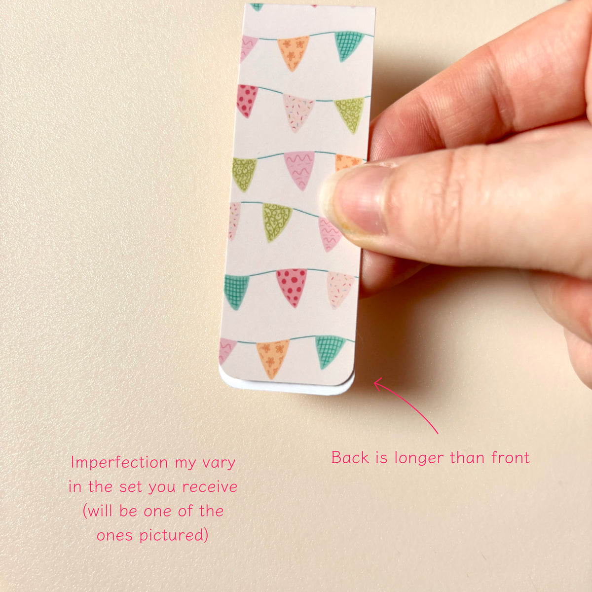 A hand holds a small magnetic bookmark with a colourful bunting pattern on a beige background. Pink text at the bottom explains that imperfections vary in the set and notes the back is longer than the front.