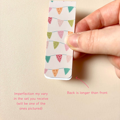 A hand holds a small magnetic bookmark with a colourful bunting pattern on a beige background. Pink text at the bottom explains that imperfections vary in the set and notes the back is longer than the front.