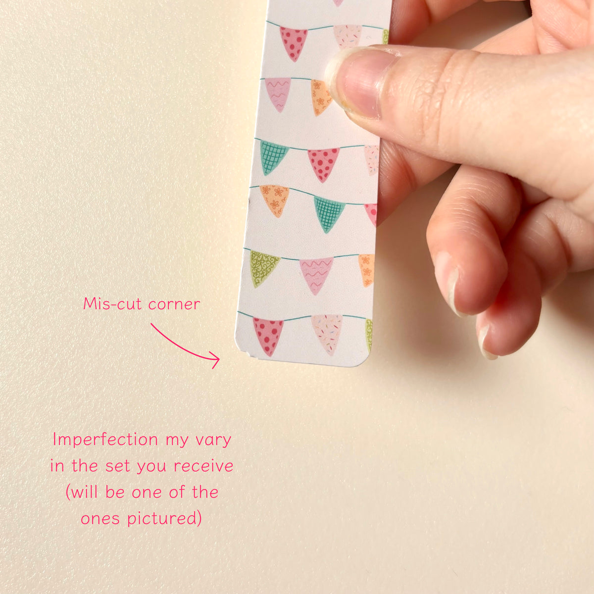 A hand holds a magnetic bookmark with a colourful bunting pattern against a plain beige background, with pink text noting a mis-cut corner and imperfection in the set.