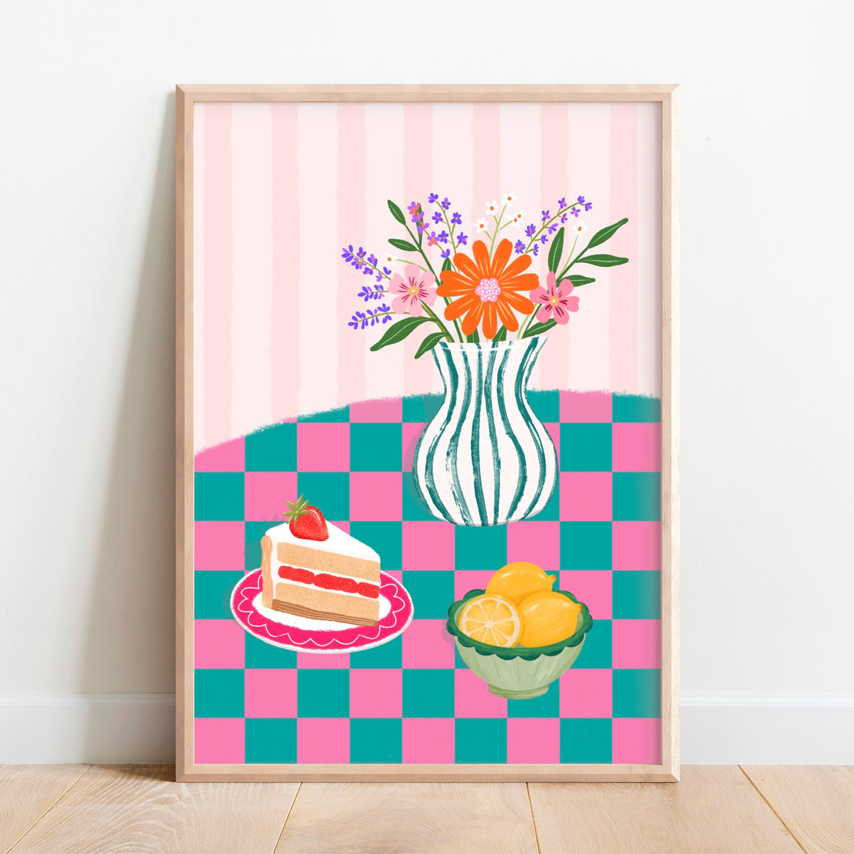 A framed art print featuring a whimsical still life illustration with a striped vase of flowers, a slice of cake on a pink plate, and lemons on a pink and teal checkered table against a pink striped background, leaning against a white wall on a light wood floor.
