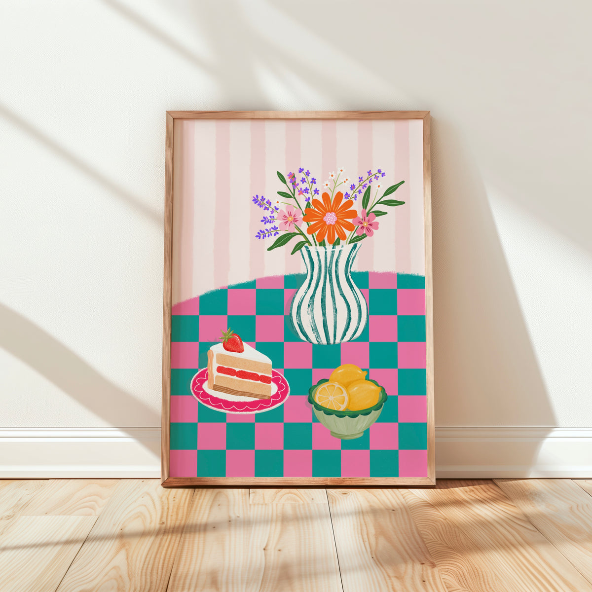 A framed art print featuring a colourful table scene with a striped pink background, a vase of flowers, a slice of cake on a plate, and a bowl of lemons, leaning against a white wall on a light wood floor.