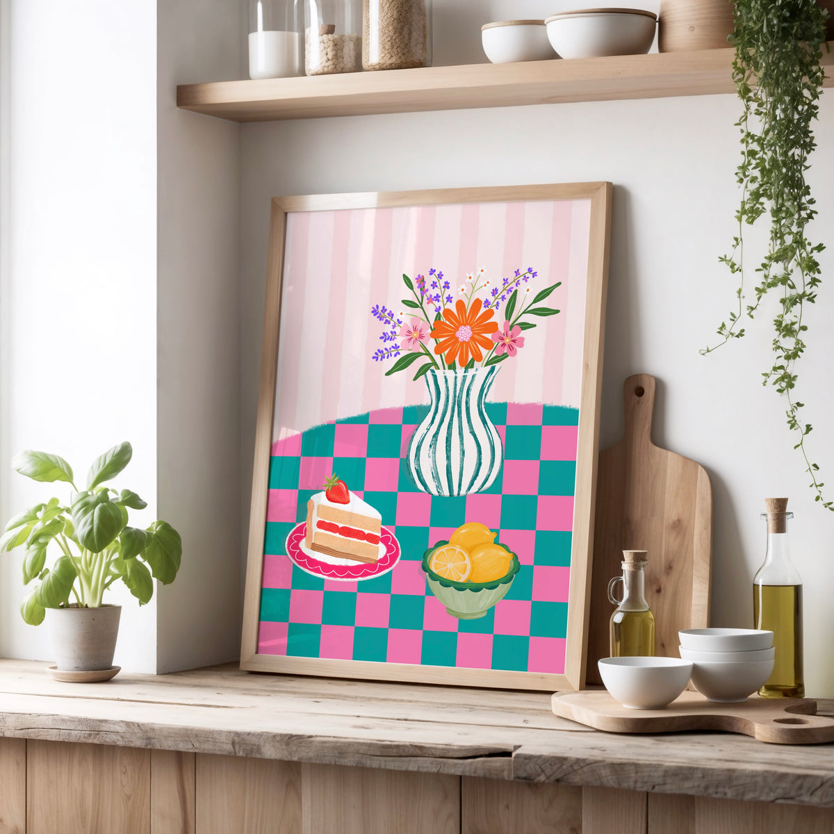 A framed art print featuring a colourful table scene with a striped pink and teal background, a striped vase of flowers, a slice of cake, and lemons, leaning on a wooden kitchen counter next to a potted plant and bowls.