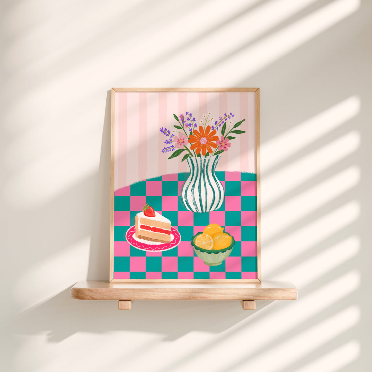 A framed art print featuring a colourful illustration of a striped vase with flowers, a slice of cake on a plate, and a bowl of lemons, displayed on a wooden easel against a sunlit cream wall.