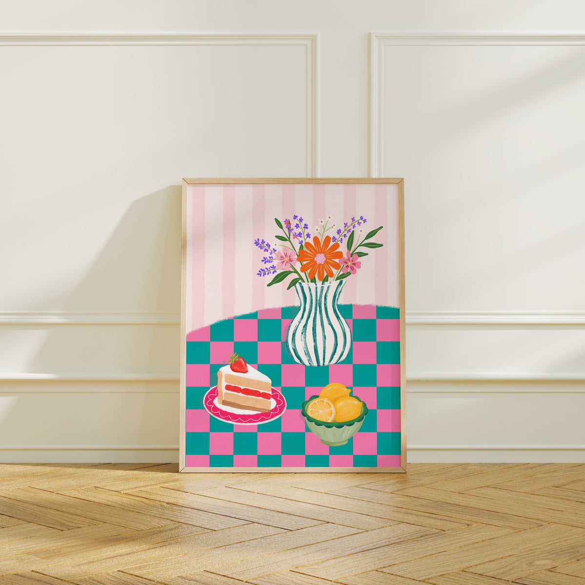 A framed art print featuring a colourful picnic scene with a striped pink-and-white background, a vase of flowers, a slice of cake, and lemons, leaning against a cream paneled wall on a wood floor.