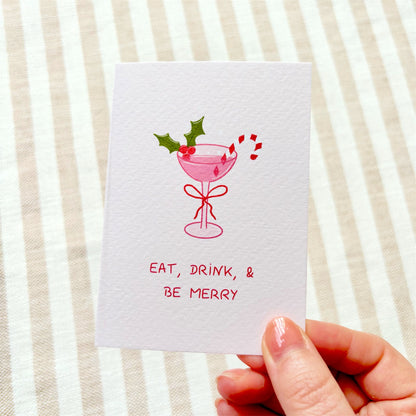 A hand holds a white greeting card with a pink cocktail illustration, green holly leaves, red berries, candy canes, and the text 'EAT, DRINK, & BE MERRY' against a beige and white striped fabric background.