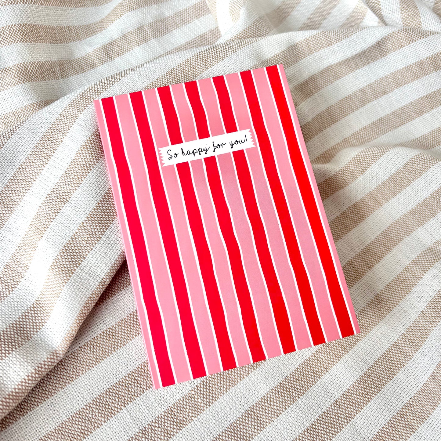 Red and pink striped card with 'So happy for you!' text against a striped fabric background.