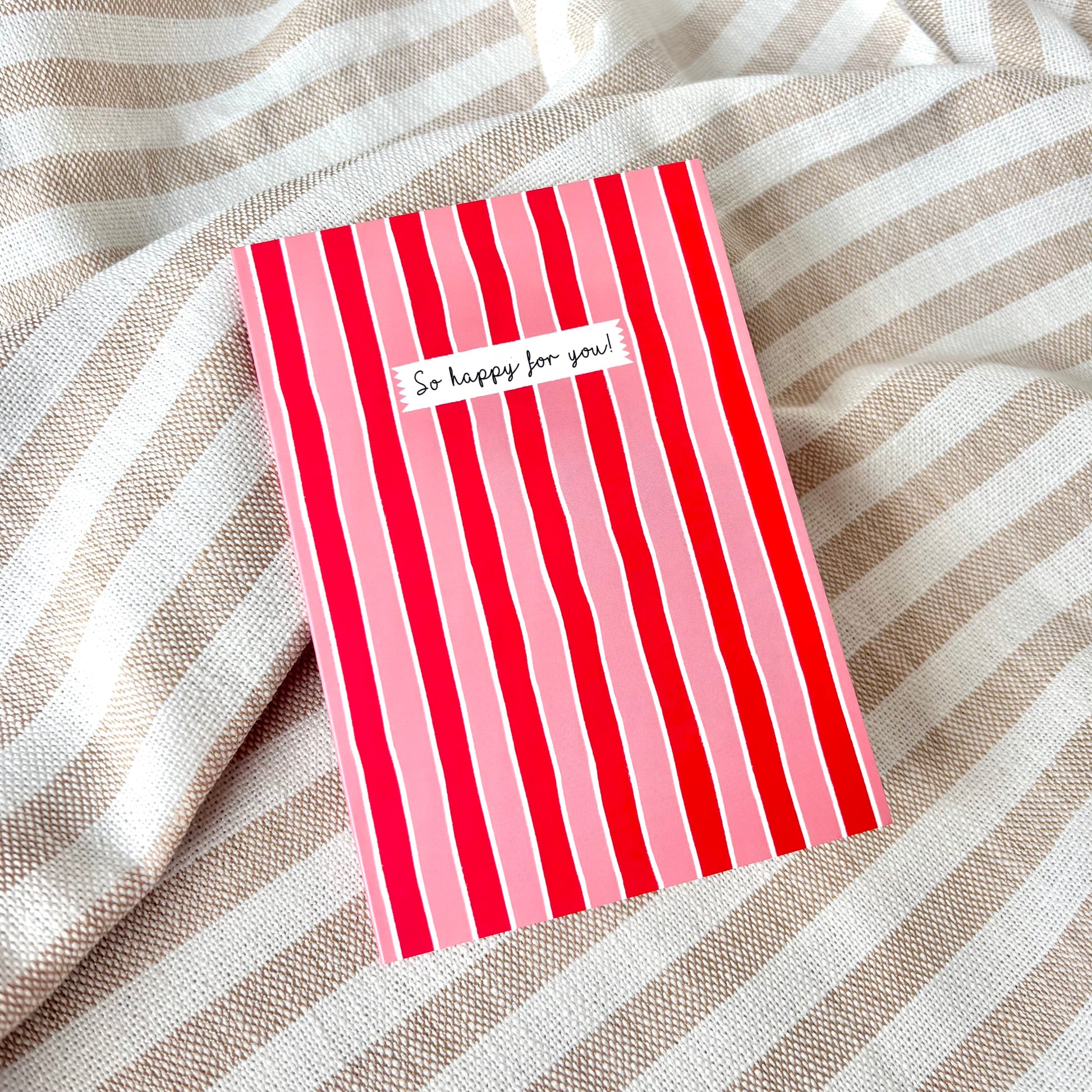 Red and pink striped card with 'So happy for you!' text against a striped fabric background.