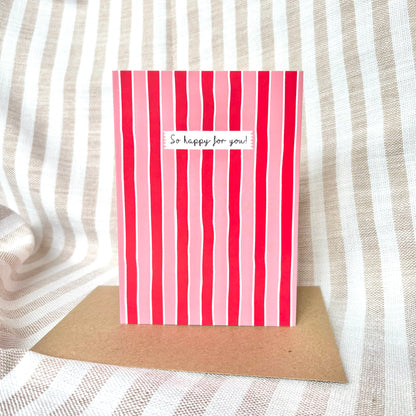 Red and pink striped card with 'So happy for you!' text on a brown kraft envelope against a striped fabric background.