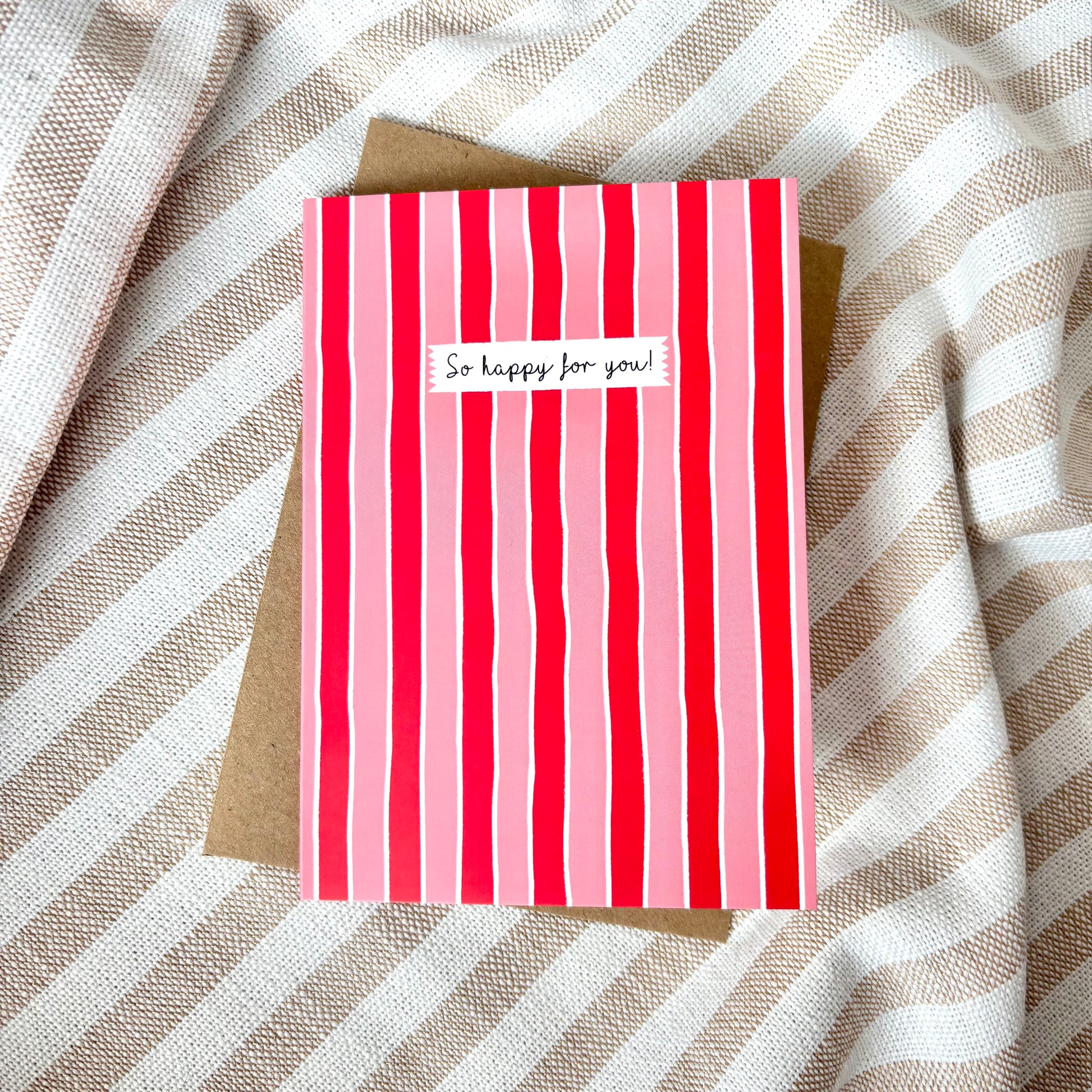 Red and pink striped card with 'So happy for you!' text on a beige envelope against a striped fabric background.