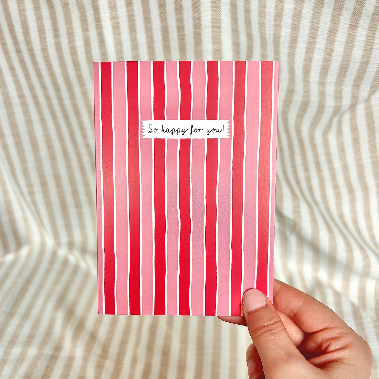 Hand holding a red and pink striped card with 'So happy for you!' text on a striped background