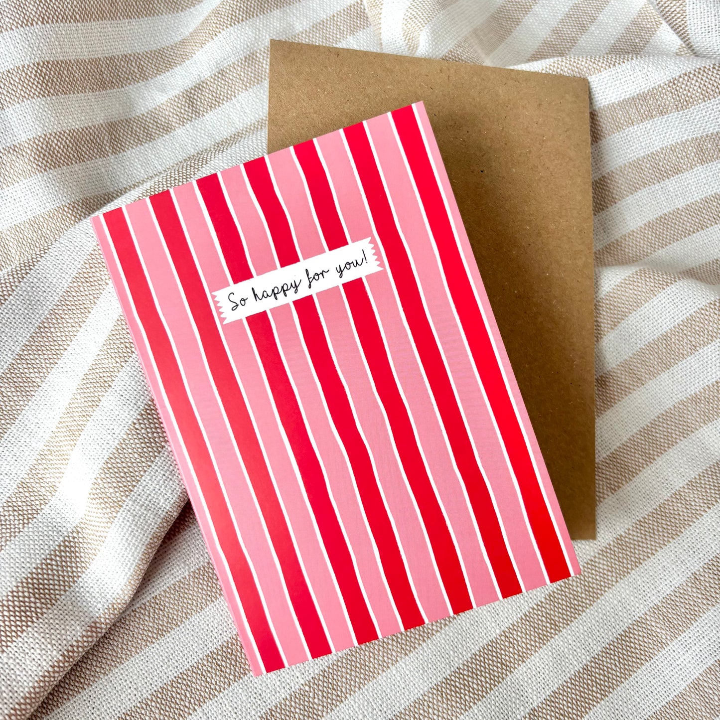 Greeting card with red and pink striped design on a brown envelope, placed on a striped fabric background.