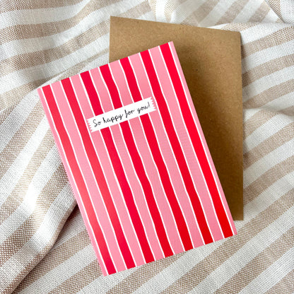 Greeting card with red and pink striped design on a brown envelope, placed on a striped fabric background.