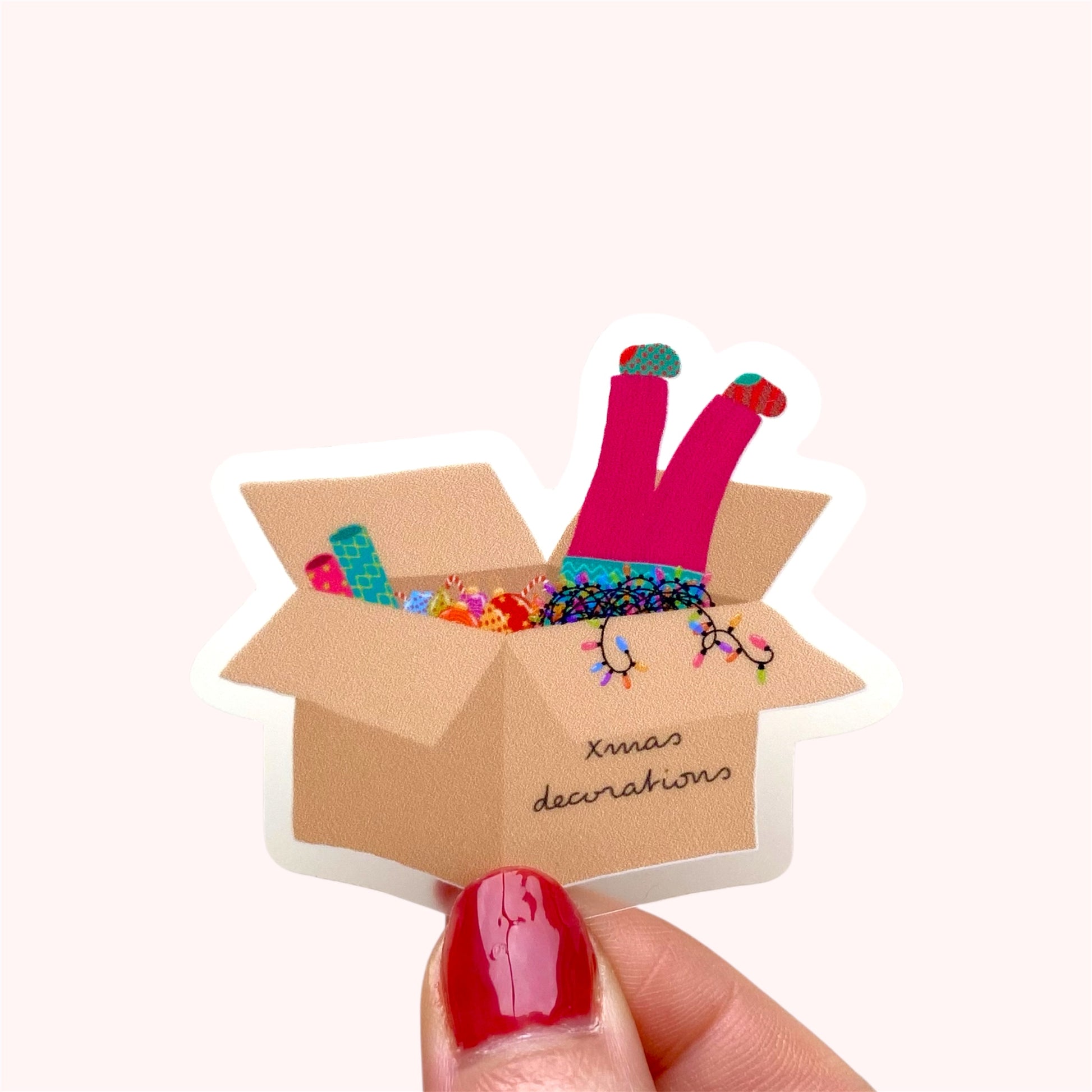 Sticker of a cardboard box labelled 'Xmas decorations' with colourful Christmas lights and ornaments and a girls legs sticking out in pink trousers in mismatched socks, held up against a white background.