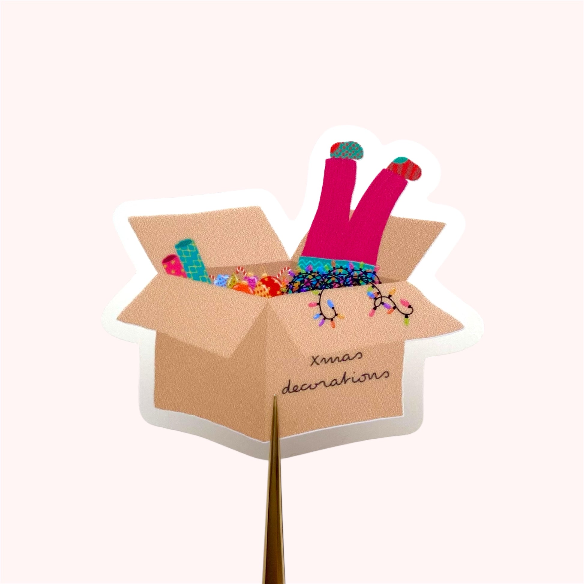 Sticker of a cardboard box with Christmas decorations and a girls legs dangling out with pink trousers and mismatched socks, on a white background