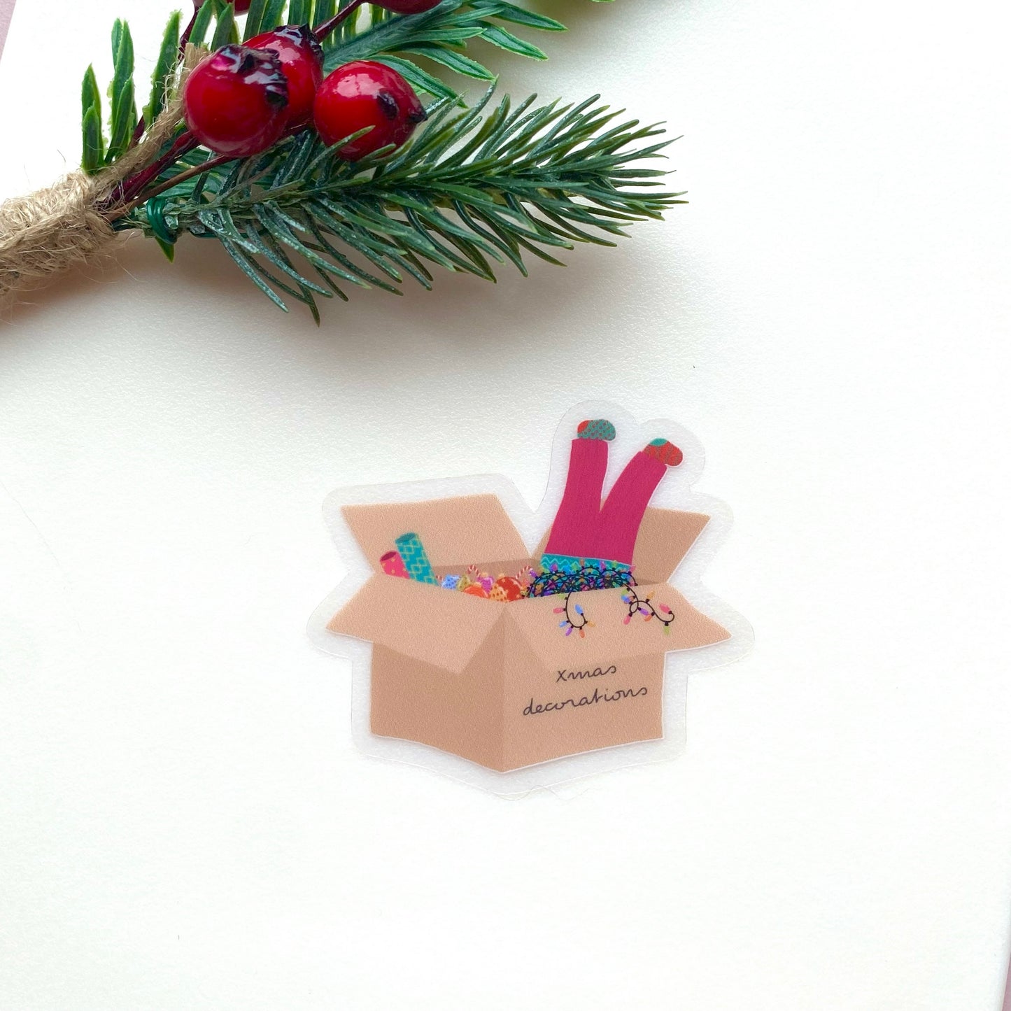 Sticker of a cardboard box with Christmas decorations and a girls legs dangling out with pink trousers and mismatched socks, on a white background with festive foliage and berries.