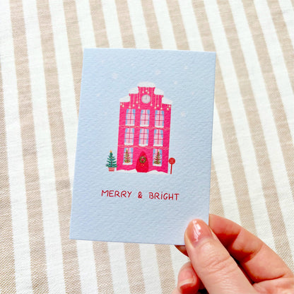 A hand holding a light blue Christmas greeting card with a pink illustration of a snow-covered Dutch house and the words 'MERRY & BRIGHT' on a beige and white striped fabric background.