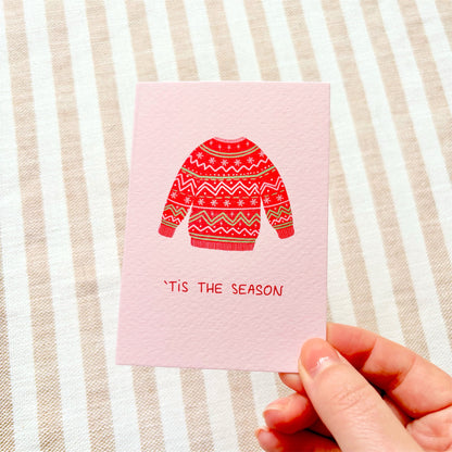 A hand holding a pink greeting card with a red, green and white patterned festive jumper illustration and the text '“Tis the Season” on a beige and white striped fabric background.