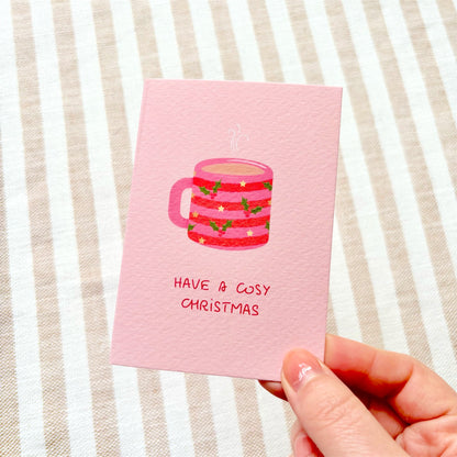 A pink Christmas greeting card held by a hand, featuring an illustration of a striped mug with holly and stars and the text 'HAVE A COSY CHRISTMAS' on a beige and white striped fabric background.