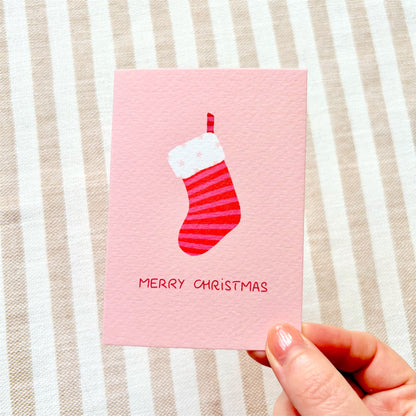 A hand holding a pink Christmas greeting card with a red and white striped stocking illustration with a starry white cuff and the words 'MERRY CHRISTMAS' in red, set against a beige and white striped fabric background.