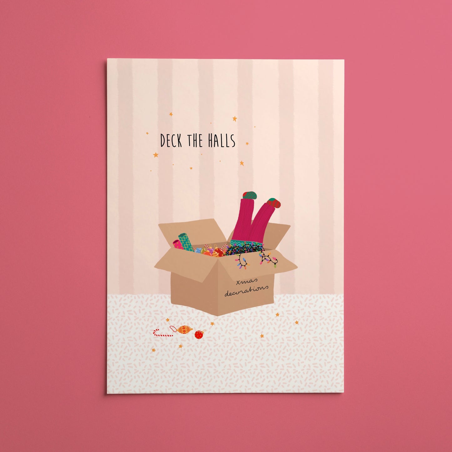 A vertical Christmas art print featuring an illustration of a girl’s legs with colourful socks popping out of a cardboard box full of decorations and string lights, with the text 'DECK THE HALLS' above it, displayed against a solid pink background.