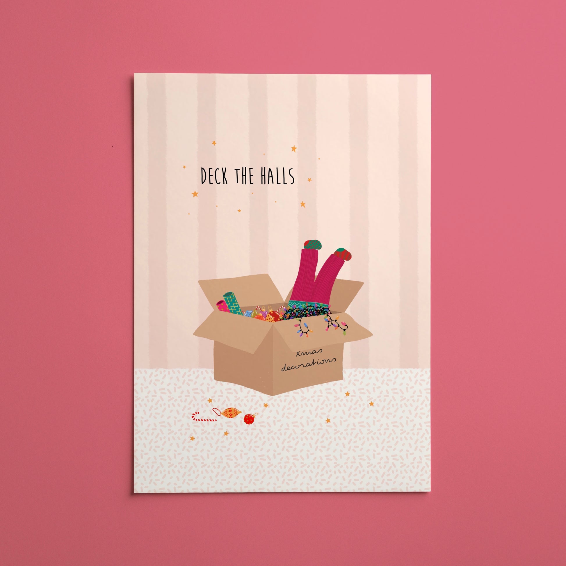 A vertical Christmas art print featuring an illustration of a girl’s legs with colourful socks popping out of a cardboard box full of decorations and string lights, with the text 'DECK THE HALLS' above it, displayed against a solid pink background.