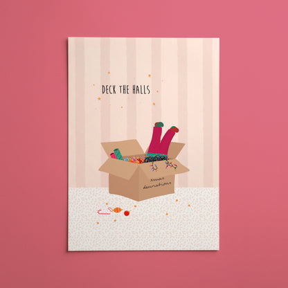 A vertical Christmas art print featuring an illustration of a girl’s legs with colourful socks popping out of a cardboard box full of decorations and string lights, with the text 'DECK THE HALLS' above it, displayed against a solid pink background.