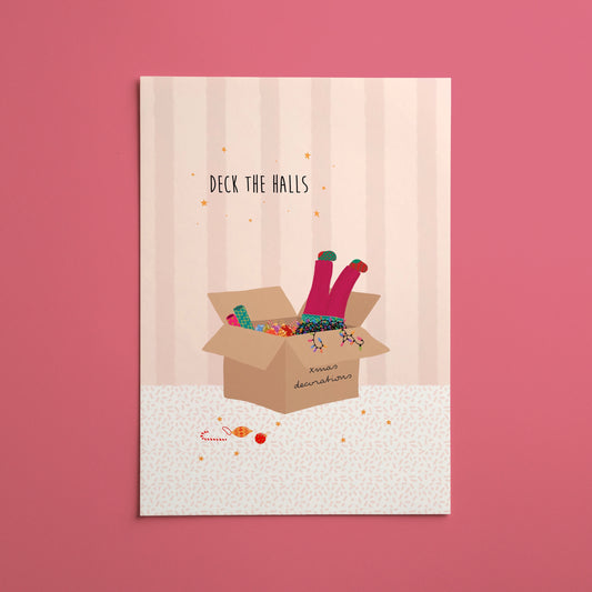A vertical Christmas art print featuring an illustration of a girl’s legs with colourful socks popping out of a cardboard box full of decorations and string lights, with the text 'DECK THE HALLS' above it, displayed against a solid pink background.