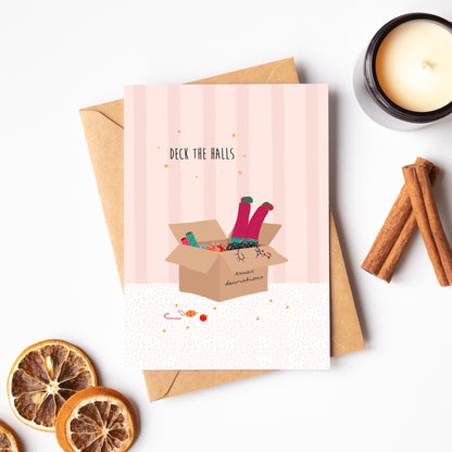 Pink striped greeting card with 'Deck the Halls' text and illustration of pink-trousered legs sticking out of a box of Christmas decorations and lights, surrounded by cinnamon sticks and dried oranges on a white background.