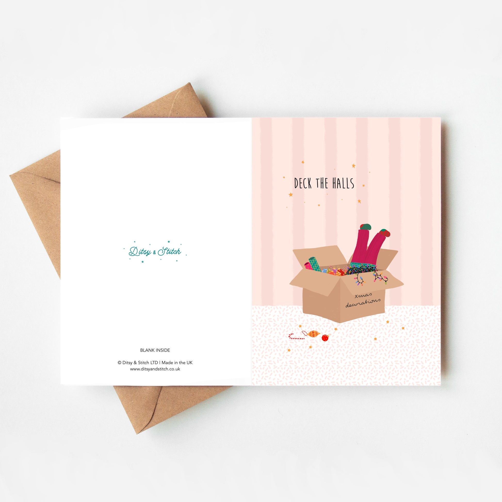 Pink striped greeting card with 'Deck the Halls' text and illustration of pink-trousered legs sticking out of a box of Christmas decorations and lights, with a brown kraft envelope
