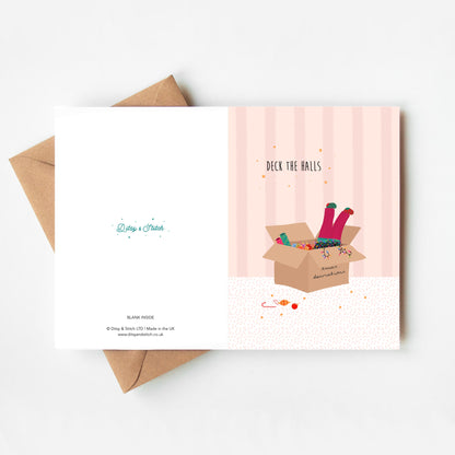 Pink striped greeting card with 'Deck the Halls' text and illustration of pink-trousered legs sticking out of a box of Christmas decorations and lights, with a brown kraft envelope