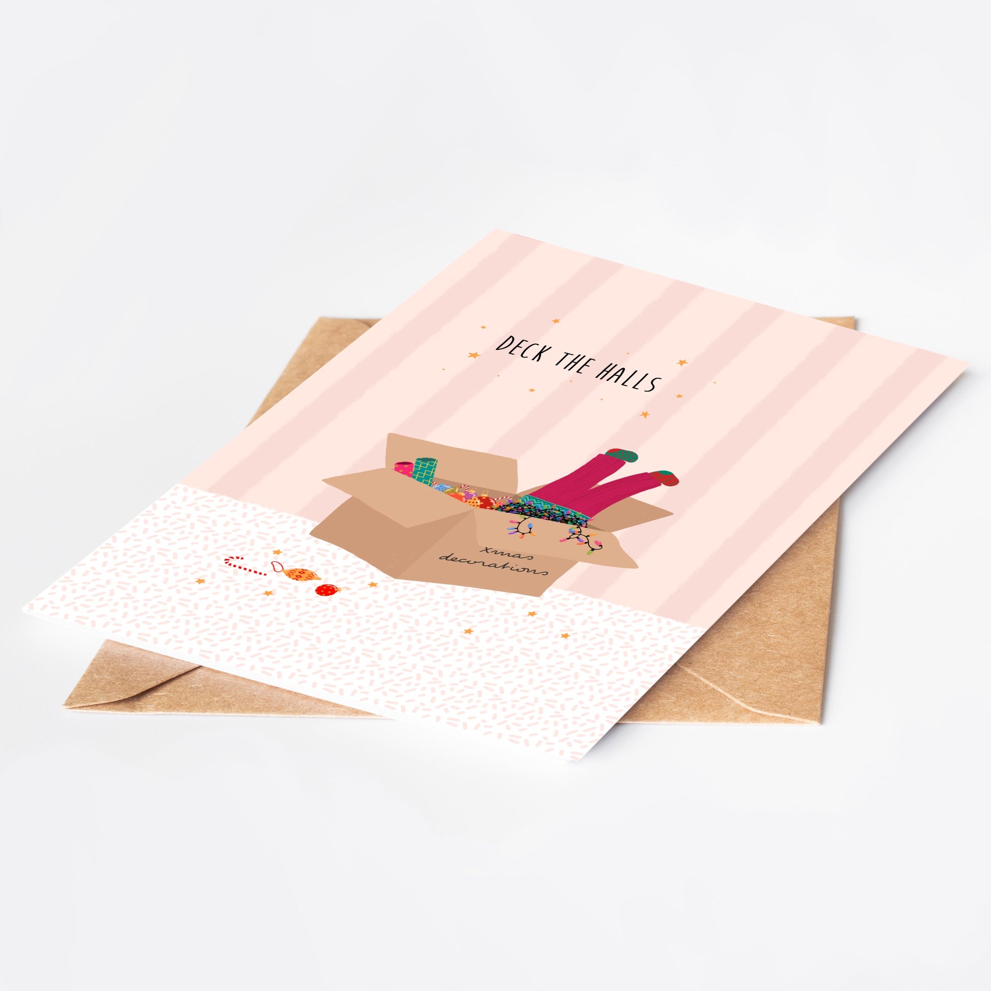 Pink striped greeting card with 'Deck the Halls' text and illustration of pink-trousered legs sticking out of a box of Christmas decorations and lights, with a brown kraft envelope