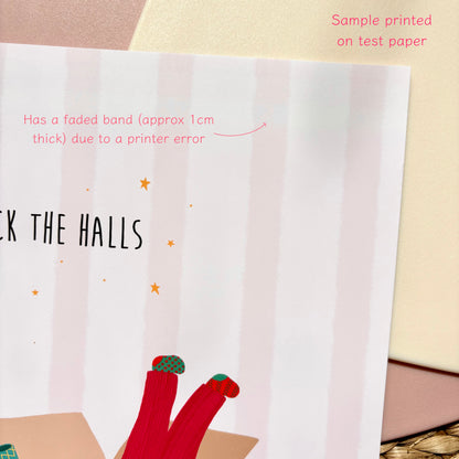 A close-up of a print sample with pale pink vertical stripes, partial black text reading 'DECK THE HALLS,' small gold stars, and a red-and-green sock illustration at the bottom. Pink overlaid text says 'Sample printed on test paper' and notes a faded band due to a printer error.